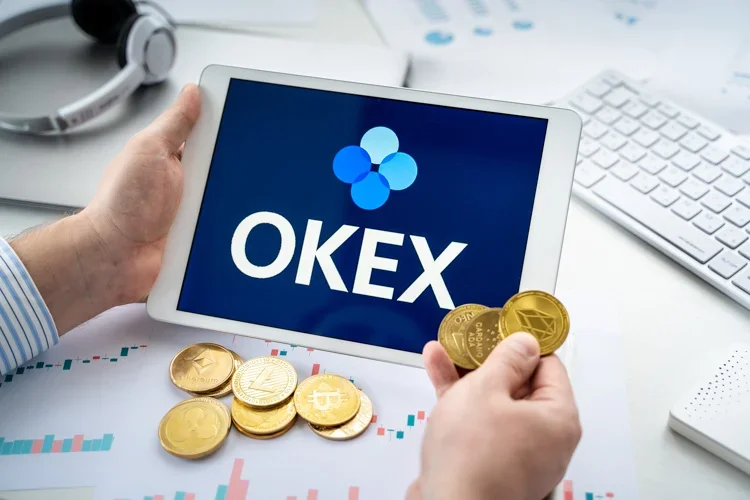 Логотип OKEx открыт на планшете и горсть монет криптовалюты в руке.