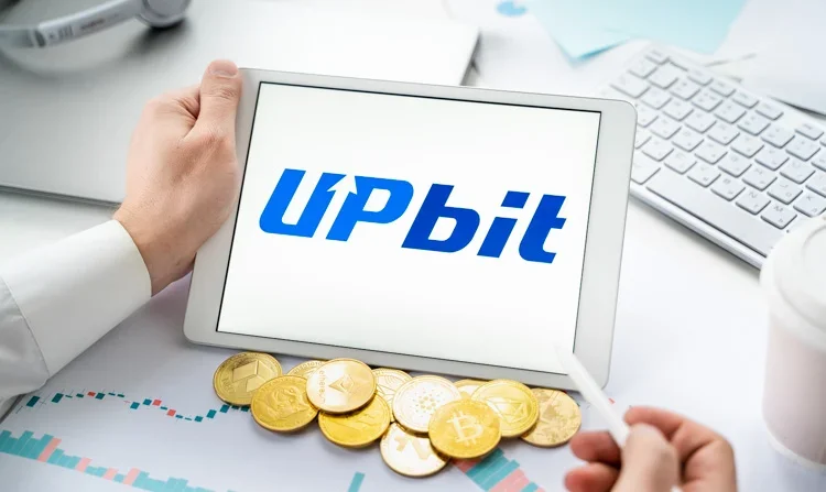 Логотип Upbit открыт на планшете и лежат монетки криптовалют.